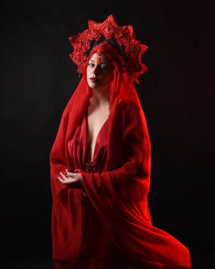 Dramatik yakın plan portre kırmızı ipek elbise giyen bir kadın model ve peçeli muhteşem bir fantezi taç. Karanlık bir stüdyonun arka planında karamsar duygusal kol hareketleriyle izole edilmiş..