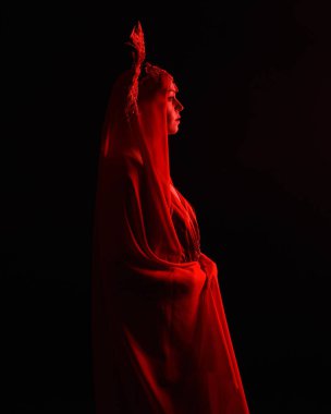 Dramatik yakın plan portre kırmızı ipek elbise giyen bir kadın model ve muhteşem bir fantezi taç ve duvak. Karanlık bir stüdyonun arka planında, siluet jant ışıklandırması ve dengesiz duygusal kol hareketleriyle izole edilmiş..