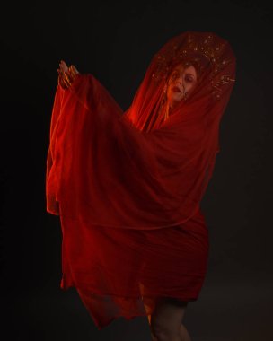 Kırmızı ipek elbise giyen, süslü altın fantezi başlık takan peçeli kadın mankenin dramatik yakın plan portresi. Huysuz, karanlık bir arka planda, siluet jant aydınlatma ve karamsar duygusal kol hareketleriyle izole edilmiş..