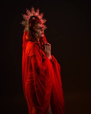 Kırmızı ipek elbise giyen, süslü altın fantezi başlık takan peçeli kadın mankenin dramatik yakın plan portresi. Huysuz, karanlık bir arka planda, siluet jant aydınlatma ve karamsar duygusal kol hareketleriyle izole edilmiş..