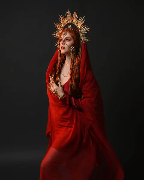 stock image dramatic close up portrait a female model wearing a red silk dress and veil robes, with a ornate golden fantasy crown headdress. isolated on a dark studio background with  silhouette rim lighting and moody emotive arm gestures.