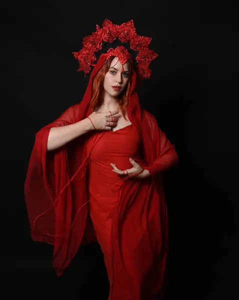 stock image close up portrait of beautiful female model wearing an ornate gothic fantasy crown and red dress. knotted string wrapped around arms, hands holding entwined rope. isolated on dark studio background.