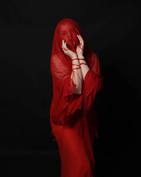 stock image close up portrait of beautiful female model wearing a veil head covering like blindfold and red dress. knotted string wrapped around arms, hands holding entwined rope. isolated on dark studio background.