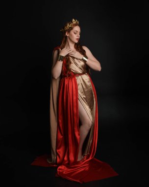Yunan kırmızı ipek Roma cübbesi giyen güzel bir kadın modelin tam boy portresi, taç takan tarihi fantezi tanrıçası kostümü. Karanlık stüdyo arka planında yalnız poz vermek.