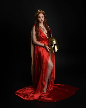 Yunan kırmızı ipek Roma cübbesi giyen güzel kadın modelin tam boy portresi, Spartalı savaşçı kaskı taşıyan tarihi fantezi tanrıçası kostümü. Karanlık stüdyo arka planında yalnız poz vermek.