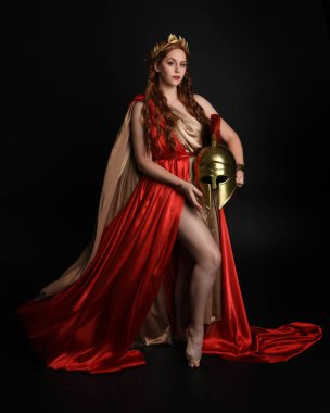 Full length portrait of beautiful female model wearing grecian red silk gold roman robes, historical fantasy goddess costume  holding spartan warrior helmet. Standing pose isolated on dark studio background. clipart