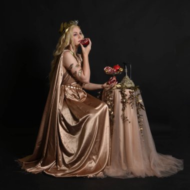 Full length portrait of beautiful female model wearing gold grecian silk toga robes, historical fantasy goddess  persephone holding pomegranate fruit. Sitting pose isolated studio background. clipart