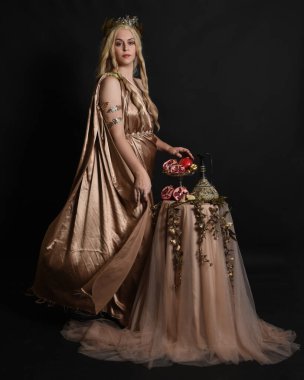 Altın sarısı ipek toga cübbe giyen güzel kadın modelin tam boy portresi, tarihi fantezi tanrıçası persephone elinde nar meyvesi tutuyor. İzole edilmiş stüdyo arkaplanı.