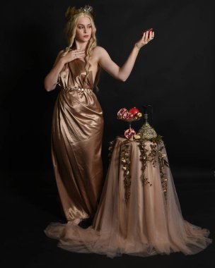 Altın sarısı ipek toga cübbe giyen güzel kadın modelin tam boy portresi, tarihi fantezi tanrıçası persephone elinde nar meyvesi tutuyor. İzole edilmiş stüdyo arkaplanı.