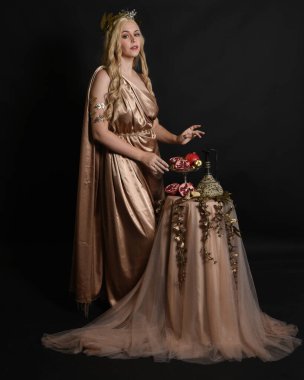 Altın sarısı ipek toga cübbe giyen güzel kadın modelin tam boy portresi, tarihi fantezi tanrıçası persephone elinde nar meyvesi tutuyor. İzole edilmiş stüdyo arkaplanı.