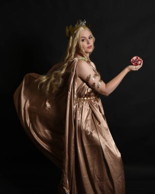 Altın sarısı ipek toga cübbe ve taç giyen güzel sarışın kadın modelin yakın plan portresi. Tarihi fantezi tanrıçası persephone elinde nar meyvesi tutuyor. Karanlık stüdyo arka planında izole.