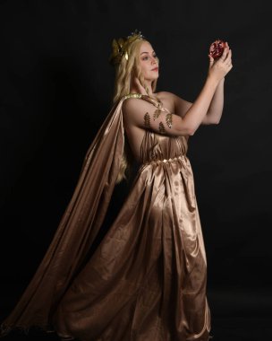 Altın sarısı ipek toga cübbe ve taç giyen güzel sarışın kadın modelin yakın plan portresi. Tarihi fantezi tanrıçası persephone elinde nar meyvesi tutuyor. Karanlık stüdyo arka planında izole.