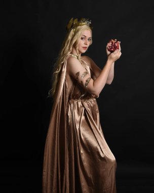 Close up portrait of beautiful blonde female model wearing gold grecian silk toga robes and crown, historical fantasy goddess persephone holding pomegranate fruit. isolated on dark studio background. clipart