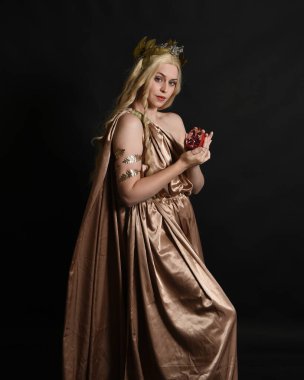 Altın sarısı ipek toga cübbe ve taç giyen güzel sarışın kadın modelin yakın plan portresi. Tarihi fantezi tanrıçası persephone elinde nar meyvesi tutuyor. Karanlık stüdyo arka planında izole.