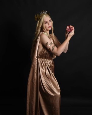 Altın sarısı ipek toga cübbe ve taç giyen güzel sarışın kadın modelin yakın plan portresi. Tarihi fantezi tanrıçası persephone elinde nar meyvesi tutuyor. Karanlık stüdyo arka planında izole.