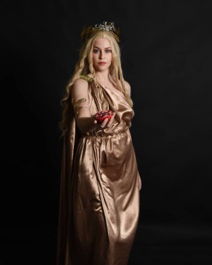 Altın sarısı ipek toga cübbe ve taç giyen güzel sarışın kadın modelin yakın plan portresi. Tarihi fantezi tanrıçası persephone elinde nar meyvesi tutuyor. Karanlık stüdyo arka planında izole.