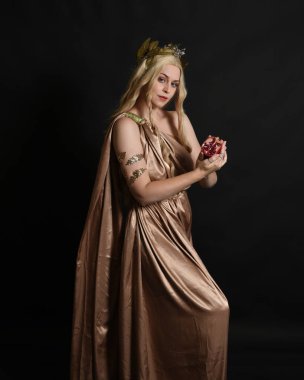 Close up portrait of beautiful blonde female model wearing gold grecian silk toga robes and crown, historical fantasy goddess persephone holding pomegranate fruit. isolated on dark studio background. clipart