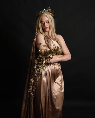 Altın sarısı ipek toga cübbe ve taç giyen güzel sarışın kadın modelin yakın plan portresi. Tarihi fantezi tanrıçası persephone elinde nar meyvesi tutuyor. Karanlık stüdyo arka planında izole.