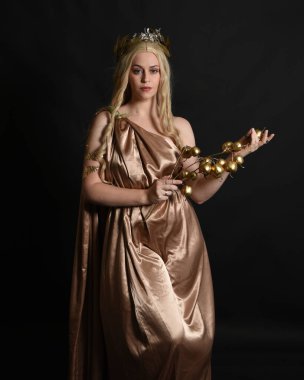 Altın sarısı ipek toga cübbe ve taç giyen güzel sarışın kadın modelin yakın plan portresi. Tarihi fantezi tanrıçası persephone elinde nar meyvesi tutuyor. Karanlık stüdyo arka planında izole.