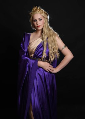 Mor ve altın renkli ipek toga cüppeli, kraliyet tacıyla, tarihi fantezi tanrıçası kostümü giymiş güzel sarışın kadın modelin tam boy portresi. Karanlık stüdyo arka planında izole edilmiş..
