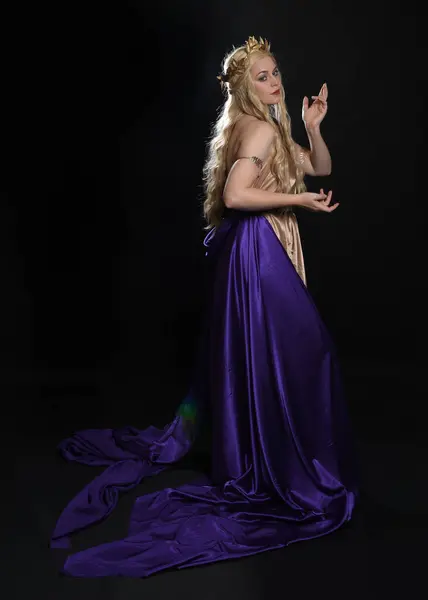 stock image full length portrait of beautiful blonde female model wearing purple and gold silken toga robes with royal crown, historical fantasy goddess costume. Isolated on dark studio background.