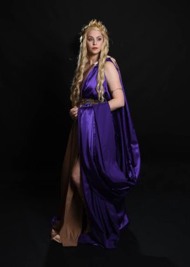 Mor ve altın renkli ipek toga cüppeli, kraliyet tacıyla, tarihi fantezi tanrıçası kostümü giymiş güzel sarışın kadın modelin tam boy portresi. Karanlık stüdyo arka planında izole edilmiş..