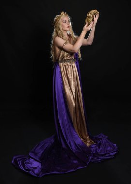 Sarışın güzel bir kadın modelin tam boy portresi mor ve altın ipek toga bornozlar ve kraliyet tacıyla, tarihi fantezi tanrıçası kostümü içinde, ritüel kafatası taşıyor. Karanlık stüdyo arka planında izole edilmiş..
