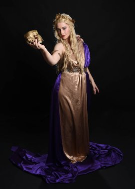 Sarışın güzel bir kadın modelin tam boy portresi mor ve altın ipek toga bornozlar ve kraliyet tacıyla, tarihi fantezi tanrıçası kostümü içinde, ritüel kafatası taşıyor. Karanlık stüdyo arka planında izole edilmiş..