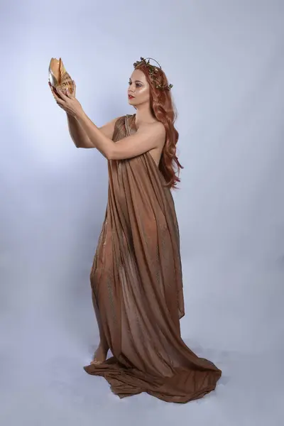 stock image Full length portrait of beautiful female model wearing silken toga robes with golden crown, historical fantasy greek goddess aphrodite costume, holding seashell skull. Isolated on studio background