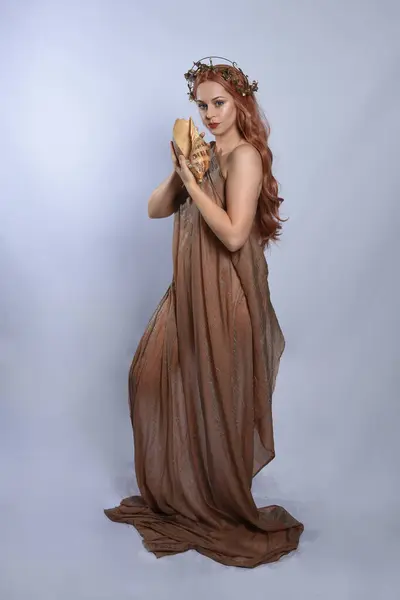 stock image Full length portrait of beautiful female model wearing silken toga robes with golden crown, historical fantasy greek goddess aphrodite costume, holding seashell skull. Isolated on studio background