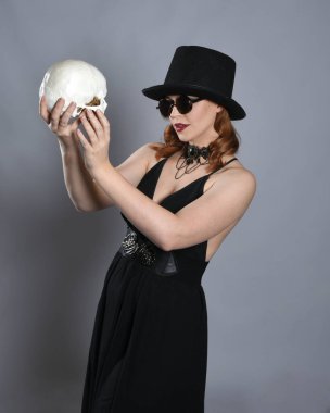 Siyah elbiseli, gözlüklü ve silindir şapkalı cadılar bayramı kostümü giymiş güzel bir kadın modelin yakın portresi. Stüdyonun arka planında izole bir figür. İskelet bir insan kafatasını tutmak.