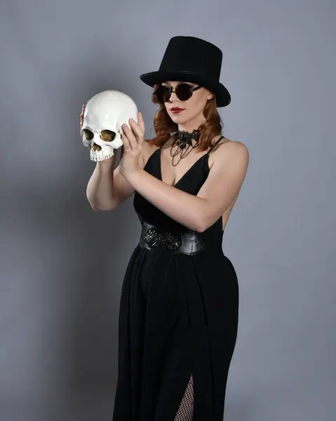 stock image Close up portrait of beautiful  female model wearing black dress and steampunk spectacles and top hat halloween costume.  Isolated figure on studio background. Holding a human skeleton skull.