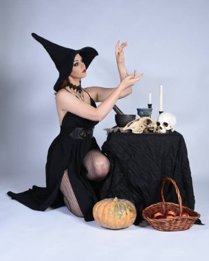  Çekici siyah cadı elbisesi giyen güzel bir kadın modelin portresi, sivri şapkalı cadılar bayramı kostümü. Okült iksir masasında sihirli döküm poz yalnız figür stüdyo arka planı