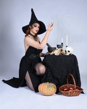  Çekici siyah cadı elbisesi giyen güzel bir kadın modelin portresi, sivri şapkalı cadılar bayramı kostümü. Okült iksir masasında sihirli döküm poz yalnız figür stüdyo arka planı