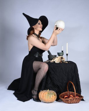  Çekici siyah cadı elbisesi giyen güzel bir kadın modelin portresi, sivri şapkalı cadılar bayramı kostümü. Okült iksir masasında sihirli döküm poz yalnız figür stüdyo arka planı