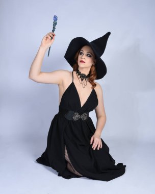 Cazibeli siyah cadı elbisesi giyip cadılar bayramı kostümü giyen güzel kızıl saçlı bir kadının tam boy portresi. Sihirli değnek tutan diz çökmüş poz, büyü yapıyor. Stüdyo arkaplanındaki izole kişi