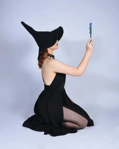 stock image Full length portrait of beautiful female red head model wearing  glamorous black witch dress with pointy hat halloween costume.  Kneeling pose  holding magical wand, casting spells.  isolated figure on studio background