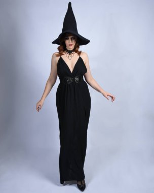 Cazibeli siyah cadı elbisesi giyip cadılar bayramı kostümü giyen güzel kızıl saçlı bir kadının tam boy portresi. Yürüyen pozlar, hareketli kollar, stüdyo arka planında izole bir figür.