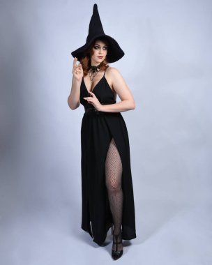 Cazibeli siyah cadı elbisesi giyip cadılar bayramı kostümü giyen güzel kızıl saçlı bir kadının tam boy portresi. Yürüyen pozlar, hareketli kollar, stüdyo arka planında izole bir figür.