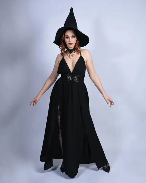 stock image Full length portrait of beautiful female red head model wearing  glamorous black witch dress with pointy hat halloween costume.   walking pose with gestural arms, isolated figure on studio background