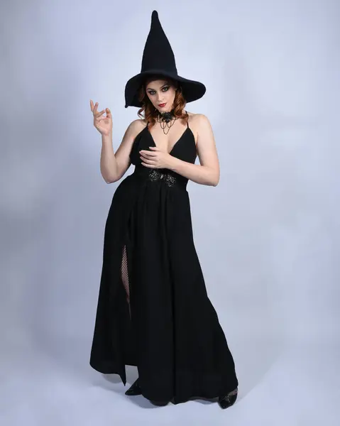 stock image Full length portrait of beautiful female red head model wearing  glamorous black witch dress with pointy hat halloween costume.   walking pose with gestural arms, isolated figure on studio background