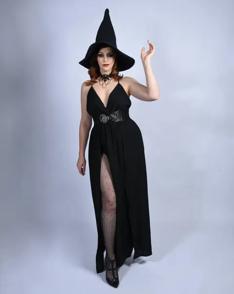 stock image Full length portrait of beautiful female red head model wearing  glamorous black witch dress with pointy hat halloween costume.   walking pose with gestural arms, isolated figure on studio background