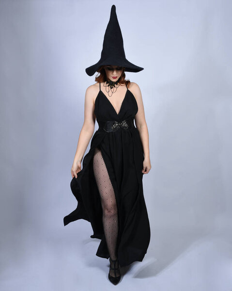 Full length portrait of beautiful female red head model wearing  glamorous black witch dress with pointy hat halloween costume.   walking pose with gestural arms, isolated figure on studio background