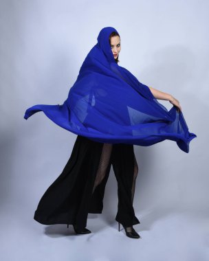 Mavi fantezi cadılar bayramı kostümlü, göz kamaştırıcı siyah cadı elbisesi giyen güzel kızıl saçlı mankenin tam boy portresi. Hareketli kollarıyla ayakta duran, stüdyo arka planında izole edilmiş bir figür.