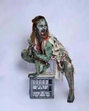 Korkunç cadılar bayramı kostümü giyen korkunç kadın modelin tam boy portresi yırtık giysiler ve sahte kan ölümsüz bir zombi karakteri gibi. Karanlık stüdyo arka planında çömelmiş bir figür.