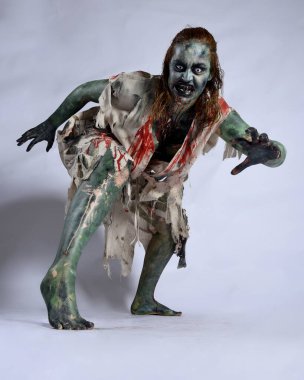 Korkunç cadılar bayramı kostümü giyen korkunç kadın modelin portresi yırtık giysiler ve sahte kan ölümsüz bir zombi karakter gibi. İzole bir figür, çömelmiş, karanlık stüdyo arkaplanı.