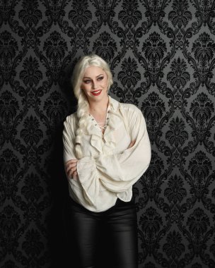 Güzel sarışın kadın modelin portresi, klasik gotik fantezi fırfırlı bluz, beyaz gömlek, deri pantolon ve uzun sarı örgülü saçlar. Siyah brokar bir duvar kağıdının üzerinde el hareketleriyle poz vermek, kasvetli bir siluet aydınlatma.