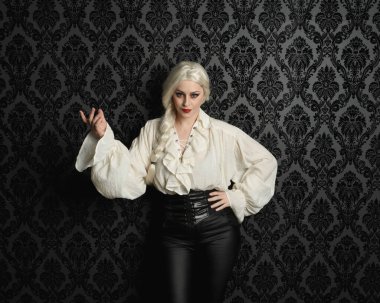 Güzel sarışın kadın modelin portresi, klasik gotik fantezi fırfırlı bluz, beyaz gömlek, deri pantolon ve uzun sarı örgülü saçlar. Siyah brokar bir duvar kağıdının üzerinde el hareketleriyle poz vermek, kasvetli bir siluet aydınlatma.