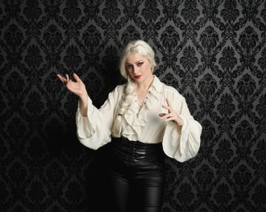 Güzel sarışın kadın modelin portresi, klasik gotik fantezi fırfırlı bluz, beyaz gömlek, deri pantolon ve uzun sarı örgülü saçlar. Siyah brokar bir duvar kağıdının üzerinde el hareketleriyle poz vermek, kasvetli bir siluet aydınlatma.