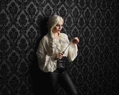 Güzel sarışın kadın modelin portresi, klasik gotik fantezi fırfırlı bluz, beyaz gömlek, deri pantolon, bıçak tutan sarı saç silahı. Huysuz siyah brokar duvar kağıdı arkaplanı